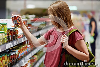 Sideways shot of pretty young femake customer holds canned goods in glass container, stands in food store, dressed in casual cloth Stock Photo