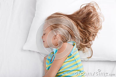 Sideways shot of small kid wears nightwear, being deep in sleep, rests at bed, lies on white bed, enjoys bedtime, rests after acti Stock Photo