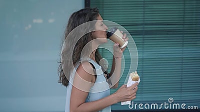 Sidosikt av den nätta unga kvinnan som äter och dricker kaffe, medan gå lager videofilmer Sidosikt av den nätta unga kvinnan som äter och dricker kaffe, medan gå lager videofilmer