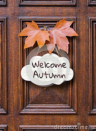 Sign with lettering words Welcome Autumn decorated with two orange maple leaves hanging on dark wooden entrance door. Stock Photo