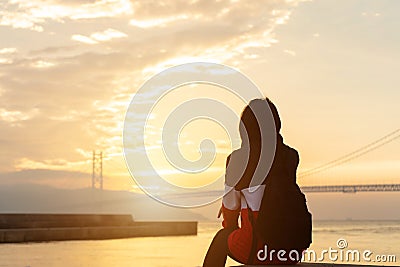 Silhouette alone young woman relax siting on riverside ocean and watching sunset and cloud sky with akashi kaikyo bridge. Stock Photo