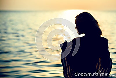 Silhouette of a beautiful woman standing with her back to the camera in the sea water at sunrise holding a large wide-brimmed hat Stock Photo