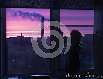 A silhouette of a child Toddler at the window looks at the pink dawn and sees smoke and urban houses Stock Photo