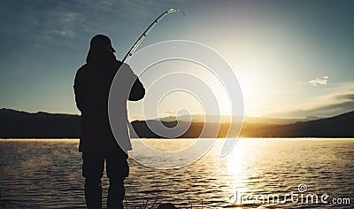 Silhouette fisherman with fishing rod at sunrise sunlight, outline man enjoy hobby sport on evening lake, person catch fish Stock Photo