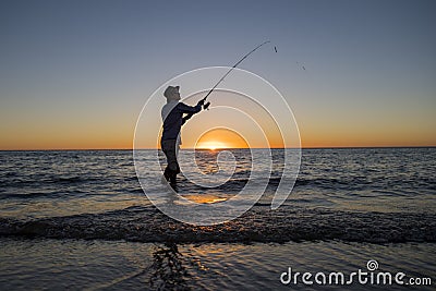 Silhouette of fisherman with hat on the beach with fish rod standing on sea water fishing at sunset with beautiful orange sky in v Stock Photo