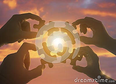 Silhouette four hands trying to connect a puzzle piece with a sunset background. Stock Photo