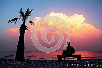 Silhouette girl with backpack sitting on bench on an empty beach with lonely palm tree and looking at an amazing purple sunset. Stock Photo