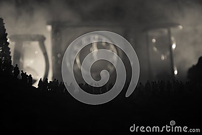 Silhouette of a large crowd of people in forest at night standing against a big arrow clock with toned light beams on foggy backgr Stock Photo