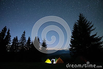 Silhouette of lonely hiker resting besides burning bonfire near illuminated tourist tents on camping site in dark Editorial Stock Photo
