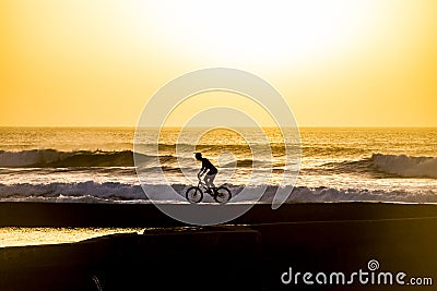 Silhouette of a male mountainbiker at sunset at the sea with wave for surfers in background - scenic place for sport adventure Stock Photo