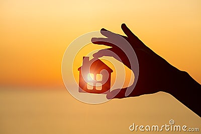 Silhouette of man`s hand holding small wooden house against sunset or sunrise light Stock Photo