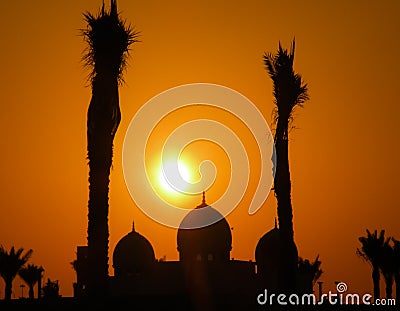 Silhouette of a Mosque with Orange Sky and sun framed by Palm Trees Stock Photo