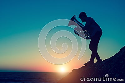 Silhouette of musician playing the tuba on rocky sea coast during amazing sunset. Stock Photo