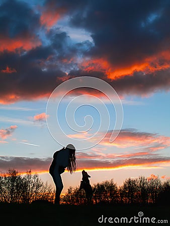 Silhouette of a teenager girl with her small yorkshire terrier in a field against beautiful sunset sky. Girl has a toy in her hand Stock Photo