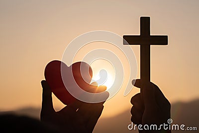 Silhouette of woman hands praying with cross and holding a red heart ball in nature sunrise background, Crucifix, Symbol of Stock Photo