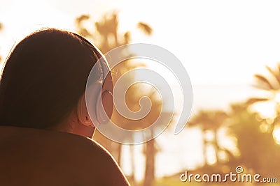 Silhouette of woman with headphones lying on the sunbed on terrace and looking on palm trees sea beach at sunset. Female relaxing. Stock Photo