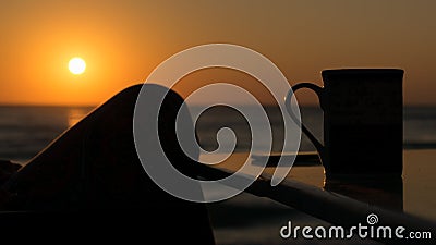 Silhouette of a woman`s foot drinking coffee in the open air at the sunrise, on the seashore Stock Photo