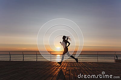 Silhouette of a young beautiful athletic girl with long blond hair in headphones, who listens to music and runs at dawn over the s Stock Photo