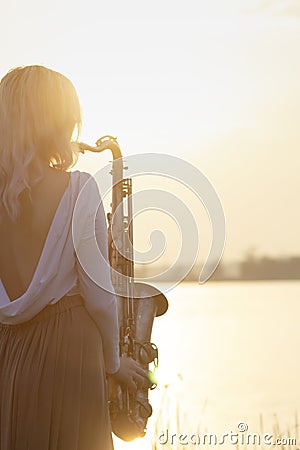 Silhouette of a young beautiful girl playing the saxophone at sunrise by the river, a woman in a long dress on the nature at Stock Photo