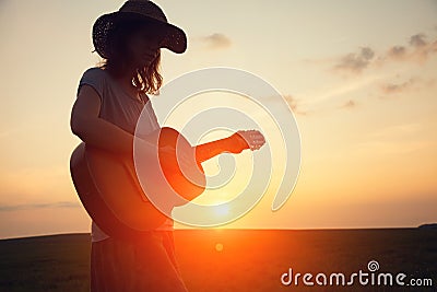 Silhouette of young free woman in straw hat playing country music on a guitar at sunset Stock Photo