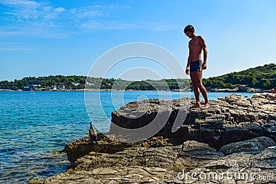 Silhouette of a young guy on a rocky Ambrela beach near the wild cormorant bird on the background of the Adriatic Sea. Pula. Stock Photo