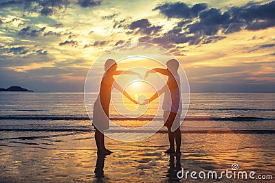 Silhouette of young romantic couple during tropical vacation, holding hands in heart shape on the ocean beach during sunset. Stock Photo