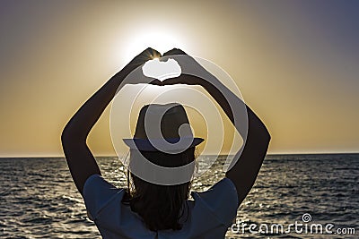 Silhouette of a young woman creating the shape of a heart with her hands with the sun nearly inside Stock Photo