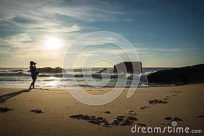 Silhouette of young woman standing on scenic sandy beach taking photos of beautiful seascape of atlantic ocean with waves in sunny Stock Photo