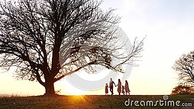 Silhouettes of happy family walking in the meadow near a big tree during sunset. Stock Photo