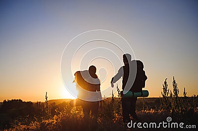 Silhouettes of two hikers with backpacks walking at sunset. Trekking and enjoying the sunset view. Stock Photo