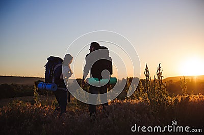 Silhouettes of two hikers with backpacks walking at sunset. Trekking and enjoying the sunset view. Stock Photo