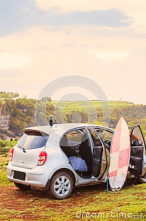 A silver grey car on a cliff with bright pink sirfboard and thermos on roof with its doors open. Surfing lifestyle on Stock Photo