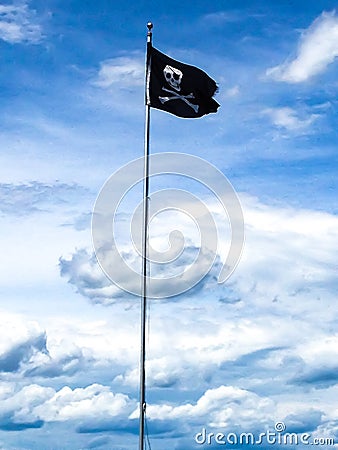 A single black skull and crossbones pirate flag waving in the air against a blue sky and clouds Stock Photo