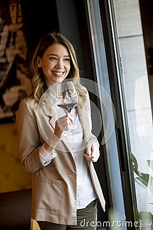 Single pretty young woman with glass of red wine standing near window and looking aside Stock Photo