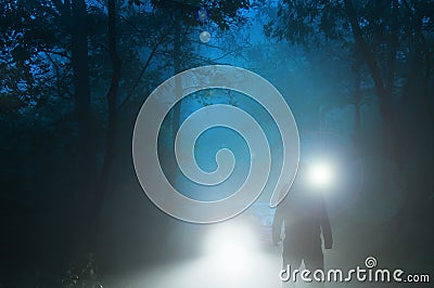 A sinister hooded figure standing in front of a car headlights on a spooky forest road on a misty evening Stock Photo