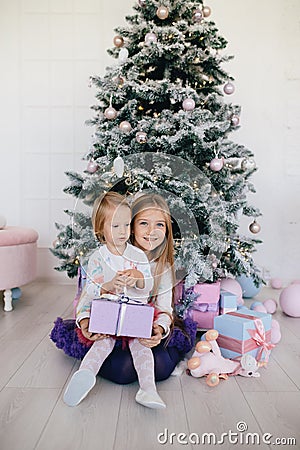 Sisters at home with Christmas tree and presents. Portrait of happy children girls with Christmas gift boxes and decorations. Stock Photo