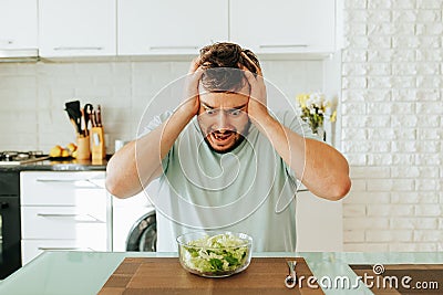 Sitting in the kitchen, a young man looks with horror at a green salad that he should eat. Stock Photo