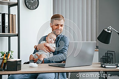 Sitting by the table with laptop. Father with toddler is indoors in the office Stock Photo