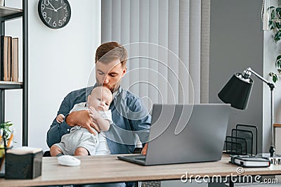 Sitting by the table with laptop. Father with toddler is indoors in the office Stock Photo