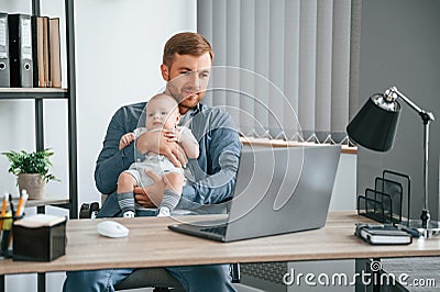 Sitting by the table with laptop. Father with toddler is indoors in the office Stock Photo