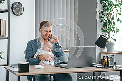 Sitting by the table with laptop. Father with toddler is indoors in the office Stock Photo
