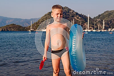 A six year old tanned boy stands holding a swimming circle and a sand shovel by the sea Stock Photo