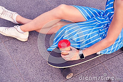 Skater woman in dress is sitting on longboard with paper cup of coffee. Close up view on hand and board. Stock Photo