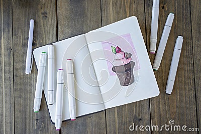 Sketching chocolate cupcake in notebook. All placed on wooden table notebook and markers. Hobby. Drawing cupcake. Top Stock Photo