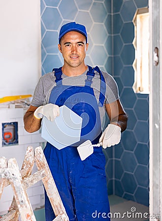 Skilled builder in work clothes posing with ceramic tiles and construction tools while repair and decoration in Stock Photo
