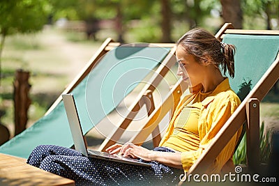 Skilled freelance woman using laptop computer while sitting on sun lounger outdoors, online working digital Stock Photo