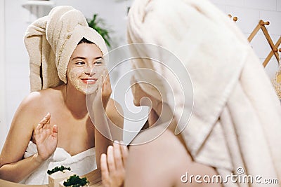 Skin Care concept. Young happy woman in towel making facial massage with organic face scrub and looking at mirror in stylish Stock Photo