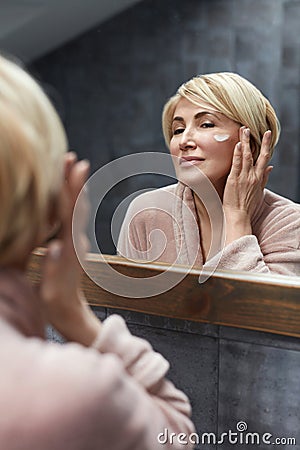 Skincare Routine. Mature Woman Uses Cosmetic Cream On Face Skin In Front Of The Mirror Reflection. Stock Photo