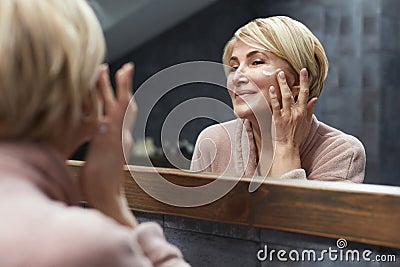 Skincare Routine. Mature Woman Uses Cosmetic Cream On Face Skin In Front Of The Mirror Reflection. Stock Photo