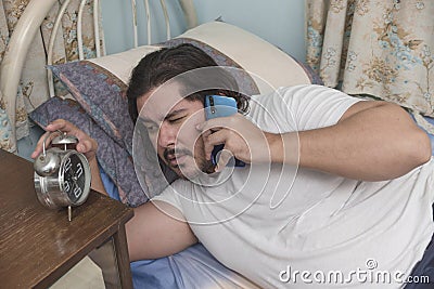 A sleepy and groggy man lying in bed checks the time on his alarm clock while talking to a client. Freelancer taking up rush work Stock Photo